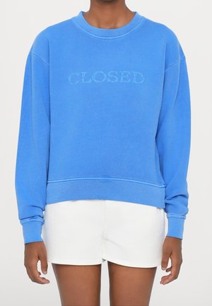 Personne portant un sweat-shirt bleu vif avec le texte "CLOSED" devant et un short blanc, sur un fond clair uni.