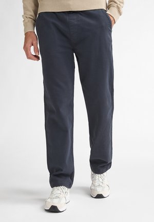 Marineblaue Baumwollhose mit entspannter Passform, ausgestattet mit Seitentaschen und Knopfverschluss. Kombiniert mit hellfarbenen Sportschuhen.