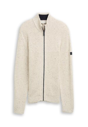 Kermanvärinen neuletakki, jossa on vetoketjukiinnitys, taitettu tekstuuri, korkea kaulus ja hienovaraisia täpliä. Mukana on musta yksityiskohta hihassa.