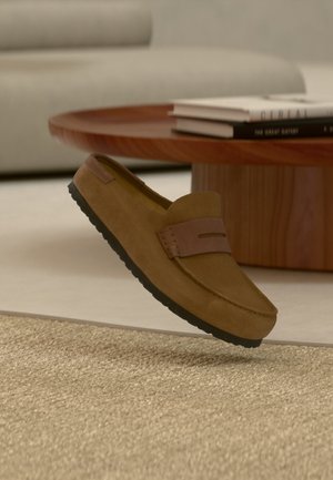 Καφέ μοκασίνι από σουέντ με μαύρη σόλα να αιωρείται πάνω από μπεζ υφαντό χαλί, ξύλινο στρογγυλό τραπέζι με στοιβαγμένα βιβλία και ανοιχτού γκρι καναπέ στο φόντο.