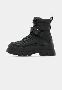 Black ankle boots with a textured upper, chunky sole, lace-up front, and adjustable strap featuring logo detail. Rubber outsole with pronounced tread.