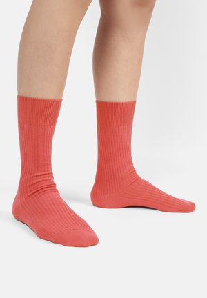 Person, die gerippte korallenfarbene Knöchelsocken trägt, steht auf hellem Hintergrund, die Füße flach auf dem Boden und nach vorne gerichtet.