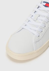 Vit sneaker med en slät läderöverdel, rund tå och platt krämfärgad gummisula. Har vita snören och en liten etikett med röda och blå accenter.