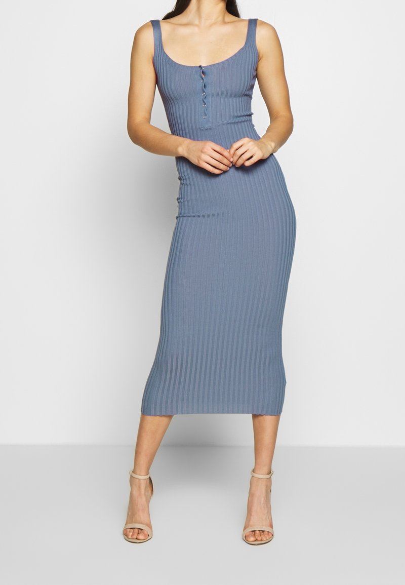 Femme portant une robe midi bleue sans manches, côtelée, avec des boutons à l'avant et des sandales à talons hauts beige, debout devant un fond uni.