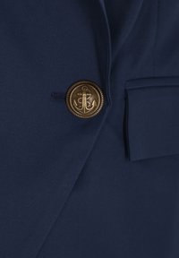 Marineblauwe stof met een getextureerde afwerking, voorzien van een messing knoop met een gegraveerd ankerontwerp. Zakdetail zichtbaar.