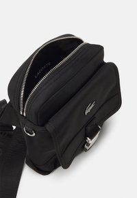 Borsa a tracolla in nylon nero con due scomparti con zipper, hardware argento, tracolla regolabile e dettaglio del logo in rilievo sulla parte anteriore.
