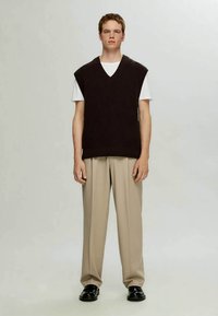 Colete de malha canelada castanho-escuro com decote em V, sobre uma t-shirt branca, combinado com calças bege e sapatos pretos. Design simples e texturizado.