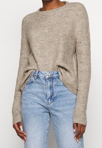 Maglione beige lavorato a maglia con scollo tondo, caratterizzato da un motivo testurizzato e spacchi laterali, abbinato a jeans a vita alta di colore azzurro chiaro.