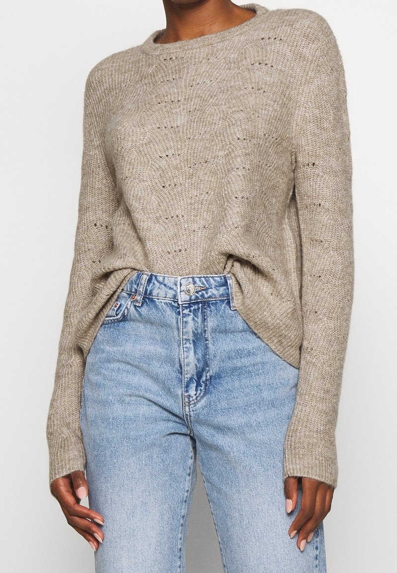 Maglione beige lavorato a maglia con scollo tondo, caratterizzato da un motivo testurizzato e spacchi laterali, abbinato a jeans a vita alta di colore azzurro chiaro.