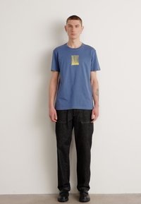 Camiseta de algodão azul com um design gráfico minimalista em amarelo e cinza, combinada com calças largas pretas texturizadas e sapatos pretos.