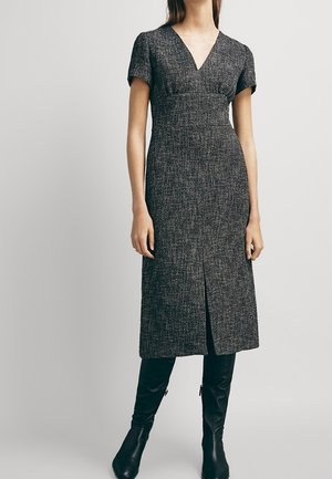 Grijs textuurjurk met een V-hals en korte mouwen. Heeft een knielange snit en een voorste split. Draagt zwarte kniehoge laarzen.