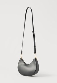 Fekete, félhold alakú bőrválltáska állítható pánttal, aranyszínű cipzárral és kapcsokkal, tiszta fehér háttér előtt.