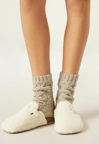 Hvide, bløde tøfler med en kabelstrikket, beige sok. Tøflerne har en rund tå og et sidespænde. Glat sål til indendørs brug.