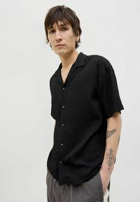 Camisa negra de manga corta con un patrón texturizado, que presenta un cierre de botones en la parte delantera y un cuello relajado. Tejido suave con un corte holgado.