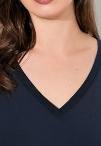 Sininen paita, jossa on V-neck-suunnittelu, valmistettu sileästä kankaasta, jossa on hienovarainen kiilto. Kauluksessa on tumman värinen reunus.