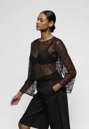 Zwarte doorschijnende blouse met borduurwerk, voorzien van een aansluitend lijfje en wijduitlopende mouwen. Gecombineerd met zwarte shorts met wijde pijpen.