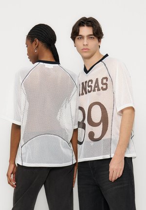 Due persone indossano maglie sportive bianche a rete; una è rivolta verso lo spettatore con la scritta "KANSAS 89" visibile, l'altra mostra la schiena, entrambe indossano pantaloni neri.