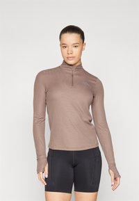 Mock neck long-sleeve top in brown, textured fabric; features a half zipper and a small blue logo. Paired with black athletic shorts.
