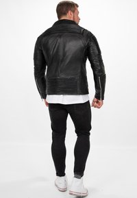 Zwarte leren jas met ribbeldetails op de schouders en rug, zilveren ritsen en een getailleerde pasvorm. Gedo wearing over een wit shirt en zwarte broek.