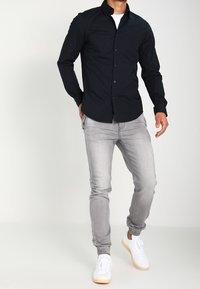 Ciemnoniebieska koszula z guzikami i klasycznym kołnierzykiem, noszona z jasnoszarymi zwężającymi się dżinsami i białymi trampkami. Gładki materiał i dopasowany krój.