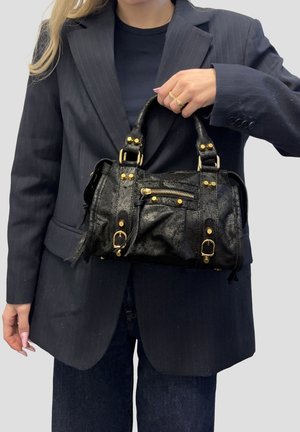 Donna con blazer e pantaloni a righe sottili scuri che tiene una piccola borsa a mano in pelle nera con dettagli dorati e tasca frontale con cerniera.