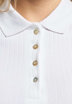 Vit topp med krage och vertikal ribbning, med fem runda knappar i ljusgrå och beige toner längs framknäppningen.