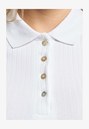 Vit topp med krage och vertikal ribbning, med fem runda knappar i ljusgrå och beige toner längs framknäppningen.