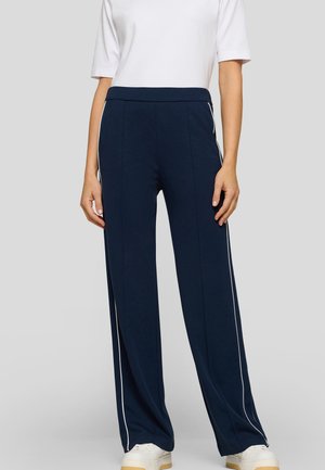 Frau trägt marineblaue, weit geschnittene Hosen mit weißen Seitenstreifen, kombiniert mit einem weißen Kurzarmoberteil und weißen Turnschuhen, stehend.