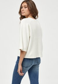 Cream-colored knit sweater with ¾ sleeves, a round neckline, and a relaxed fit. Paired with dark blue fitted jeans. Smooth texture.