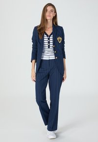 Navy pinstripe suit with gold buttons and crest, paired with a white and navy striped top. Model wears white sneakers.