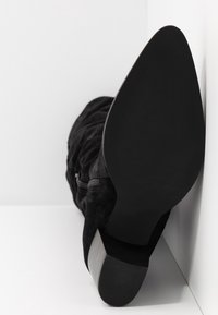 Bottines en suède noir avec un talon pointu, détail de fermeture éclair et une semelle texturée noire ; le talon a une finition lisse.