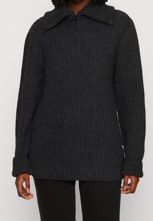 Osoba ubrana w ciemnoszary prążkowany sweter dzianinowy z krótkim, przednim zamkiem błyskawicznym i podwiniętymi mankietami, w połączeniu z czarnymi spodniami.