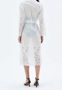 Biała sukienka z długim rękawem i paskiem w talii, zdobiona misternymi haftowanymi wzorami na rękawach i na dole. Teksturowany, lekki materiał.