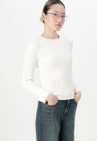 Jonge vrouw met een bril, gekleed in een witte geribbelde lange mouwtop en een spijkerbroek, met haar handen in de zakken, voor een witte achtergrond.