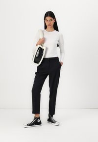 Chemise blanche à manches longues, pantalon noir ajusté, baskets montantes noires, portant un sac noir et blanc avec une bordure en polaire. Design simple.