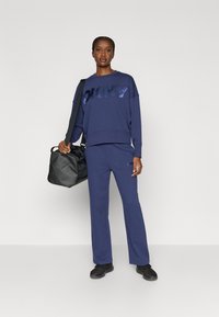 Granatowy komplet bluzy i spodni z logo DKNY w błyszczącym wykończeniu. Model nosi czarną sportową torbę typu duffle i czarne sneakersy.