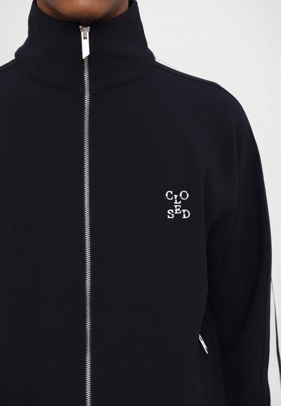 Veste zippée noire avec un col montant, une fermeture éclair argentée et un logo brodé blanc sur la poitrine gauche. Design simple et uni.