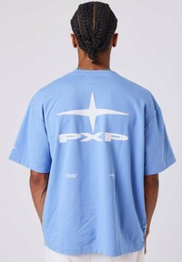 Hombre con pelo trenzado que lleva una camiseta azul clara de gran tamaño con los logotipos blancos "PXP" y estrellas en la parte trasera, de pie contra un fondo liso.