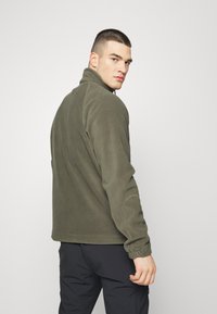 Olive green fleece jacket with a high collar, slightly fitted design, elastic cuffs, and flatlock seams. Worn over black pants.