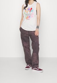 Witte mouwloze top met kleurrijke abstracte print en logo, gecombineerd met losvallende paarse tie-dye broek en kastanjebruine sneakers.