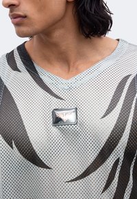 Gros plan d'une personne portant un t-shirt en maille claire avec des motifs noirs en forme de flammes et un badge rectangulaire présentant un dessin ailé sur la poitrine.