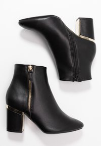 Botas de tobillo de cuero negro con punta afilada, que cuentan con un zipper lateral y acentos en tono dorado en el tacón cuadrado apilado. Textura suave.