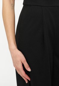 Αριστερό χέρι και χέρι να ακουμπάνε στο πλάι, φορώντας μαύρο παντελόνι με ψηλή μέση, με ένα μικρό τατουάζ στον πάνω βραχίονα.