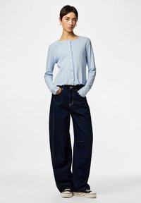 Γυναίκα που φοράει ανοιχτό μπλε ζακέτα με κουμπιά, σκούρα τζιν με φαρδιά παντελόνια και τα χέρια στις τσέπες, καθώς επίσης και μαύρα και λευκά αθλητικά παπούτσια, στέκεται μπροστά σε ένα απλό φόντο.