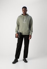 Haut à capuche vert olive avec poche avant et logo blanc, associé à un pantalon noir droit et des chaussures noires à lacets, tenant une pochette noire.