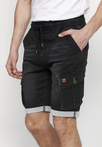 Pantalones cortos de carga negros con dobladillos enrollados, confeccionados en denim. Incluyen bolsillos con botones y detalles de costura visibles.