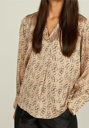 Blusa beige de manga larga con patrón circular negro, usada por una persona con cabello largo y castaño, combinada con pantalones negros.