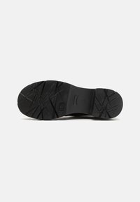 Black rubber shoe sole with a textured grip pattern, featuring zigzag lines and a rounded toe. Solid, durable design for traction.