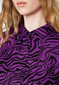 Blouse violette avec un motif audacieux de rayures zebres noires, un col pointu et des boutons de fermeture. Tissu lisse avec une légère brillance.