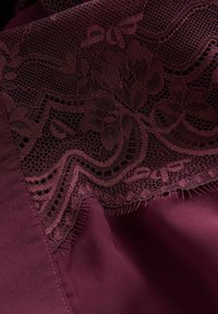 Sovrapposizione di pizzo bordeaux con motivi floreali su una base di tessuto morbido. Texture liscia con cuciture intricate lungo i bordi.